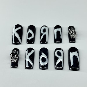 Op de afbeelding: Zwarte en witte kunstnagels. De nagels zijn zwart met witte letters die "KORN" spellen. Twee nagels hebben zilveren skelet hand bedels. De nagels zijn coffin-vormig.