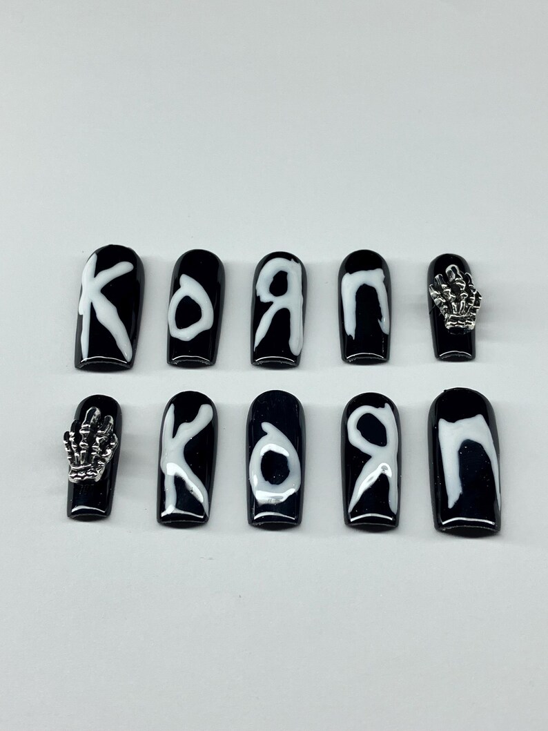 Op de afbeelding: Zwarte en witte kunstnagels. De nagels zijn zwart met witte letters die "KORN" spellen. Twee nagels hebben zilveren skelet hand bedels. De nagels zijn coffin-vormig.