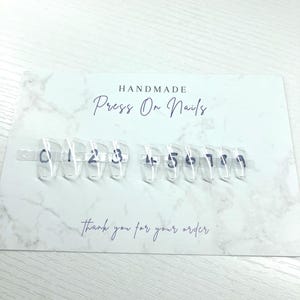 Op de afbeelding: Transparante press-on nagelmonsters met de nummers 0-9 geschreven in blauwe inkt. De nagels worden weergegeven op een witte kaart met de woorden "HANDMADE Press On Nails" en "Thank you for your order" in een scriptlettertype.