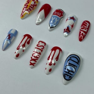 May include: A set of eleven artificial fingernails with various designs. The nails feature white, red, blue, and black colors, with text and graphic elements. Designs include dripping blood, stars, and the word "lost."