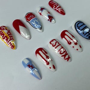 May include: A collection of ten long, almond-shaped artificial nails. The nails feature various designs, including red and white patterns, text like "Miss Sunshine" and "EST 19XX", and blue accents. The nails are arranged on a white surface.
