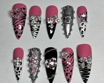 May include: Set of ten pink and white press-on nails with different designs. Each nail features a different embellishment, including a heart, a snake, a star, a cross, and a bow.