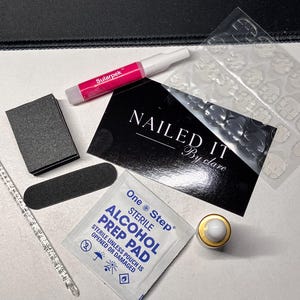 Op de afbeelding: Een verzameling nagelverzorgingsproducten, waaronder nagellijm, een nagelvijl, een nagelbuffer, een cuticle pusher, nagellijmstrips en een kleine fles. De tekst "NAILED IT" is zichtbaar.