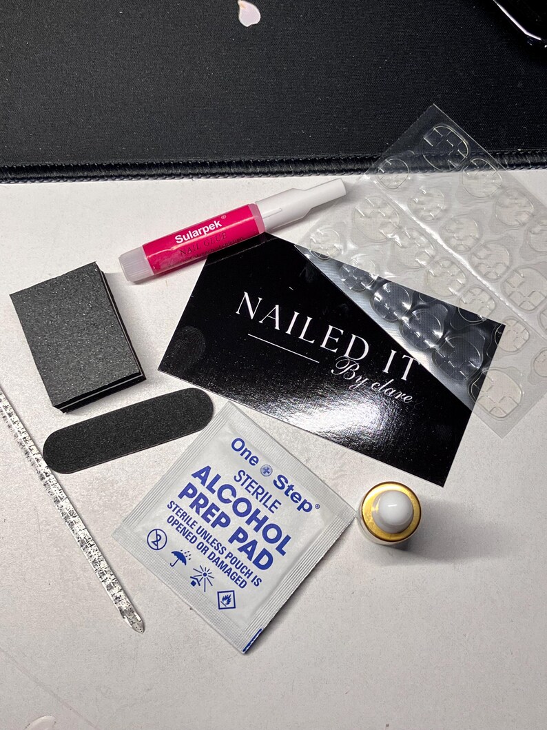Op de afbeelding: Een verzameling nagelverzorgingsproducten, waaronder nagellijm, een nagelvijl, een nagelbuffer, een cuticle pusher, nagellijmstrips en een kleine fles. De tekst "NAILED IT" is zichtbaar.