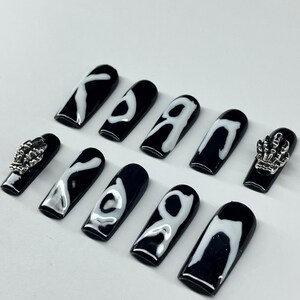 Op de afbeelding: Zwarte en witte doodskistvormige kunstnagels. De nagels hebben een glanzende zwarte basis met witte abstracte ontwerpen. Twee nagels hebben zilveren skelet hand bedels. Een set stijlvolle, draagbare kunstnagels.