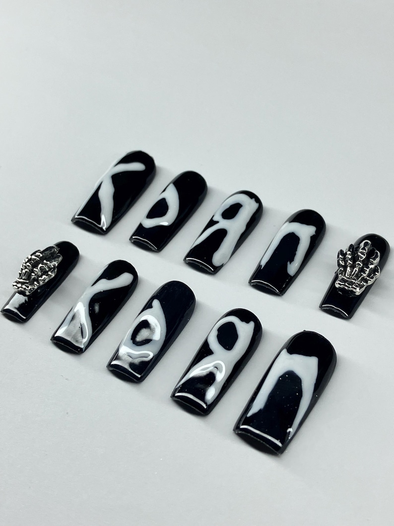 Op de afbeelding: Zwarte en witte doodskistvormige kunstnagels. De nagels hebben een glanzende zwarte basis met witte abstracte ontwerpen. Twee nagels hebben zilveren skelet hand bedels. Een set stijlvolle, draagbare kunstnagels.