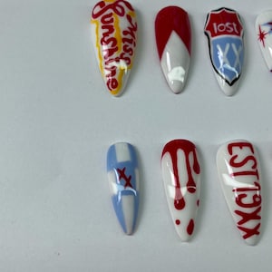 May include: A collection of artificial nails with various designs. Some nails feature text like "Sunshine" and "Lost", while others have red and white patterns, including drips and crosses. The nails are oval-shaped and set against a white background.