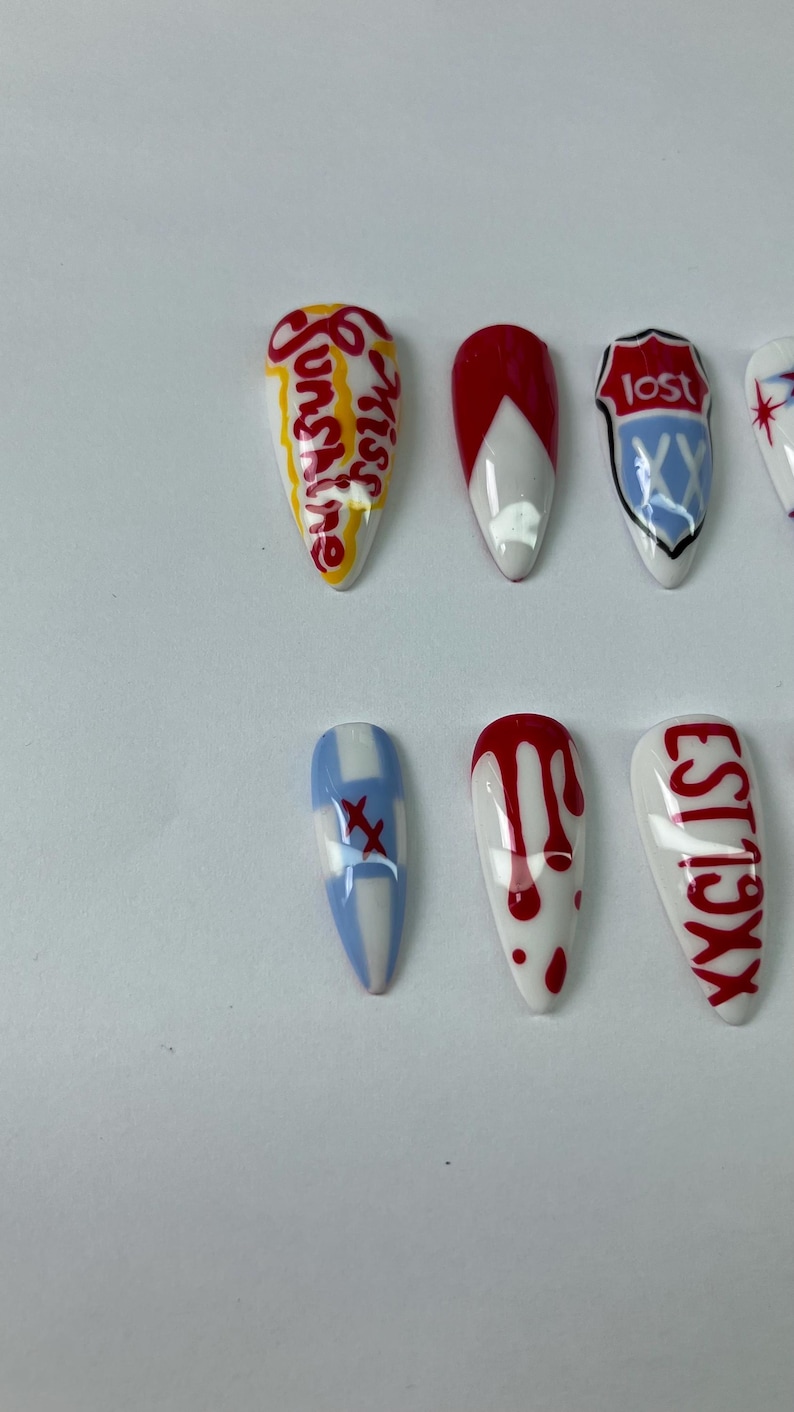 May include: A collection of artificial nails with various designs. Some nails feature text like "Sunshine" and "Lost", while others have red and white patterns, including drips and crosses. The nails are oval-shaped and set against a white background.