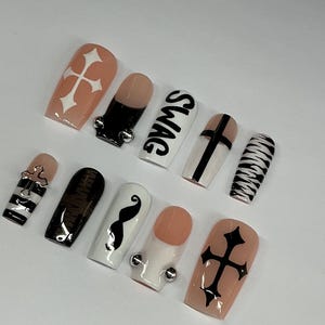 May include: A set of ten artificial nails with various designs. The nails feature a mix of colors, including black, white, and nude, with accents like crosses, stripes, and a mustache. One nail has the word "SWAG" in black.