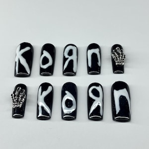 Op de afbeelding: Zwarte opdruknagels met witte letters die "KORN" spellen. Twee nagels hebben zilveren skelet hand bedels. De nagels zijn ovaal van vorm en hebben een glanzende afwerking.