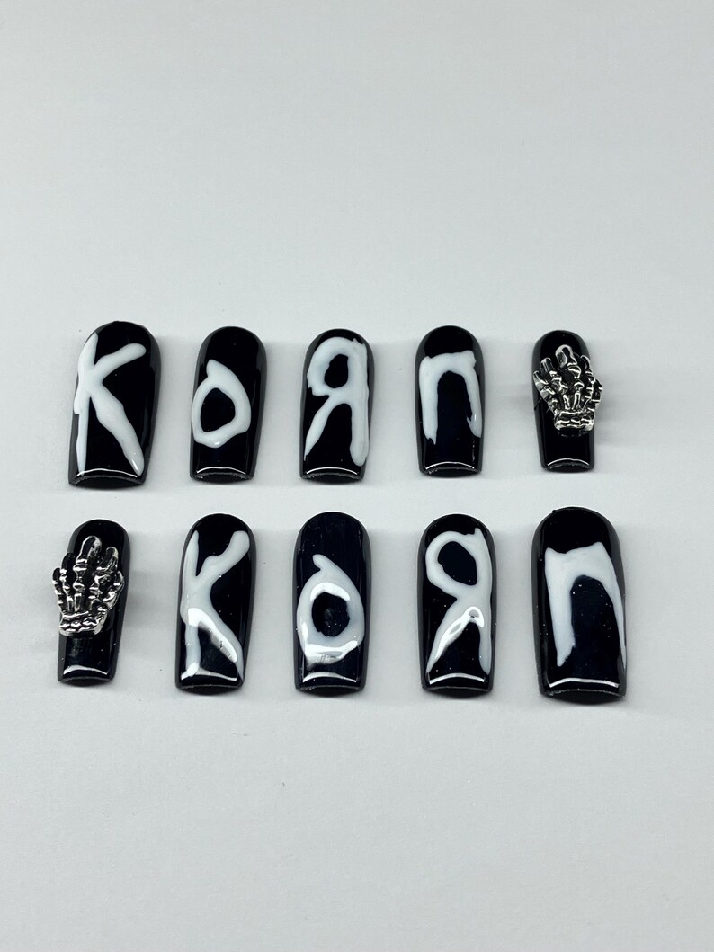 Op de afbeelding: Zwarte opdruknagels met witte letters die "KORN" spellen. Twee nagels hebben zilveren skelet hand bedels. De nagels zijn ovaal van vorm en hebben een glanzende afwerking.