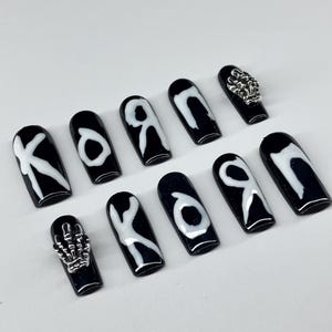 Op de afbeelding: Zwarte persnagels met witte letters en zilveren versieringen. De nagels hebben een glanzende afwerking en een doodskistvorm. Sommige nagels hebben de letters "K", "O" of "R" in het wit. Twee nagels hebben zilveren skelet hand- en schedelbedels.