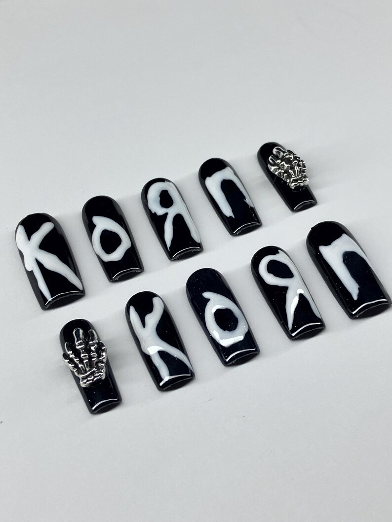 Op de afbeelding: Zwarte persnagels met witte letters en zilveren versieringen. De nagels hebben een glanzende afwerking en een doodskistvorm. Sommige nagels hebben de letters "K", "O" of "R" in het wit. Twee nagels hebben zilveren skelet hand- en schedelbedels.