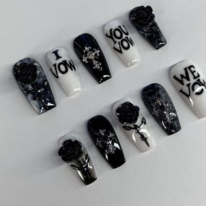 May include: A set of coffin-shaped false nails in black and white. Some nails feature a rose design, a cross, or the words "I Vow", "You Vow", and "We Vow". The nails have a glossy finish and are arranged on a white surface.