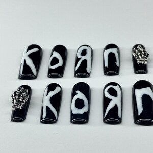 Op de afbeelding: Zwarte en witte kunstnagels met het woord "KORN" in witte letters. Twee nagels hebben zilveren skelet hand bedels. De nagels zijn glanzend en hebben een doodskistvorm.