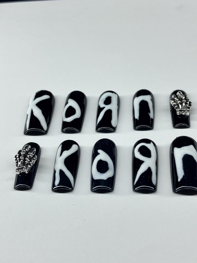 Op de afbeelding: Zwarte en witte kunstnagels met het woord "KORN" in witte letters. Twee nagels hebben zilveren skelet hand bedels. De nagels zijn glanzend en hebben een doodskistvorm.