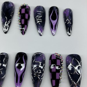 Puede incluir: Un conjunto de diez uñas artificiales largas y puntiagudas. Los diseños incluyen patrones de tablero de ajedrez, motivos de llamas y remolinos abstractos en negro, morado y plateado. Algunas uñas tienen detalles de pedrería y texto.