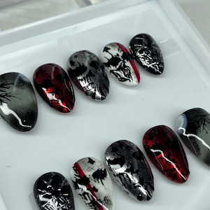 May include: A collection of artificial nails with a gothic design. The nails display skull motifs, abstract patterns, and a combination of black, red, white, and silver hues. The oval-shaped nails are presented on a white surface.