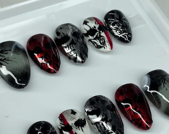 Puede incluir: Un conjunto de uñas postizas con temática gótica. Las uñas presentan una variedad de diseños, incluyendo calaveras, patrones abstractos y una mezcla de colores negro, rojo, blanco y plateado. Las uñas son de forma ovalada y están dispuestas sobre una superficie blanca.