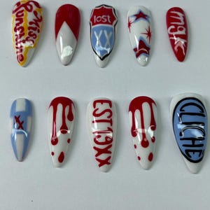 May include: A set of ten almond-shaped artificial nails. Each nail is painted with unique designs, including text like "Sunshine", "Lost", and "Est19XX", along with abstract patterns in red, white, and blue. The nails are arranged on a white surface.