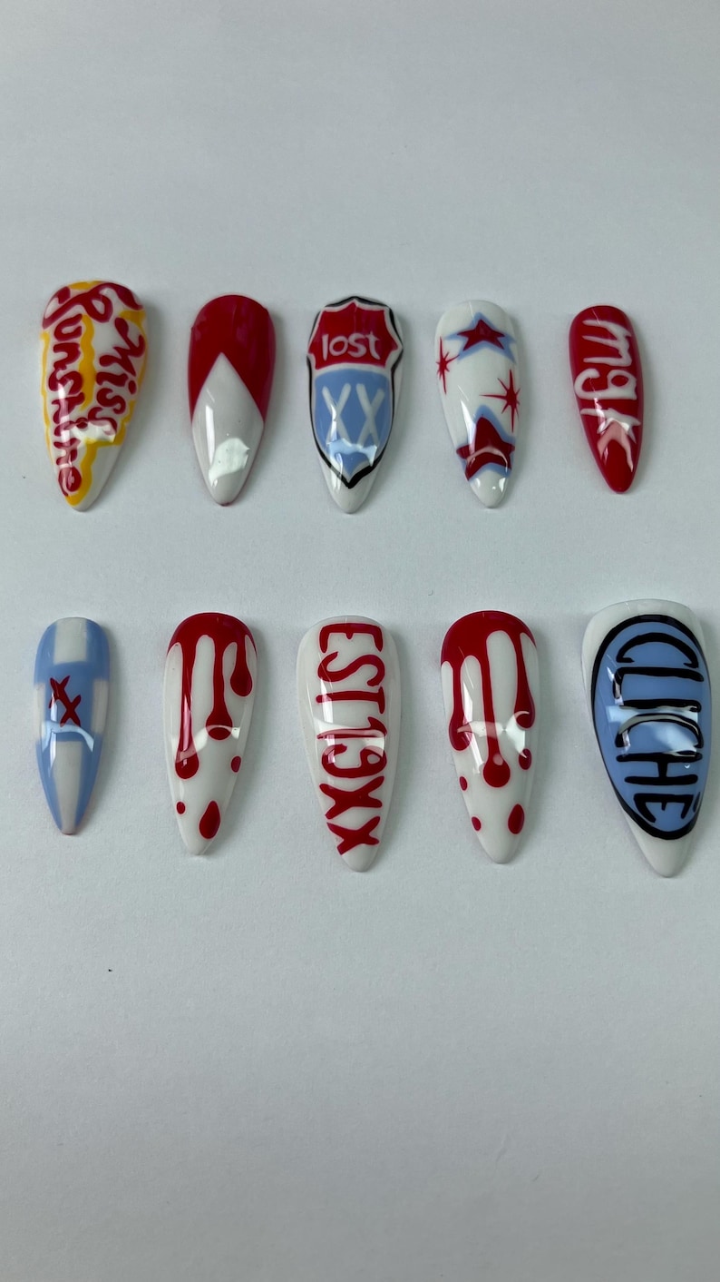 May include: A set of ten almond-shaped artificial nails. Each nail is painted with unique designs, including text like "Sunshine", "Lost", and "Est19XX", along with abstract patterns in red, white, and blue. The nails are arranged on a white surface.