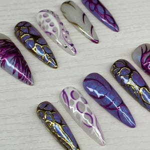 May include: A collection of long, pointed artificial nails in various designs. The nails feature shades of purple, white, and gold, with intricate patterns and embellishments. Some nails have a gold metallic edge.