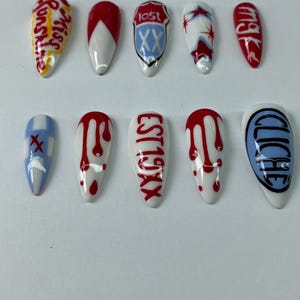 May include: A set of ten artificial nails with various designs. Some nails feature text like "Miss Sunshine" and "EST 19XX". Others have red and white patterns, including dripping blood effects and a blue and white design with the word "Lost".