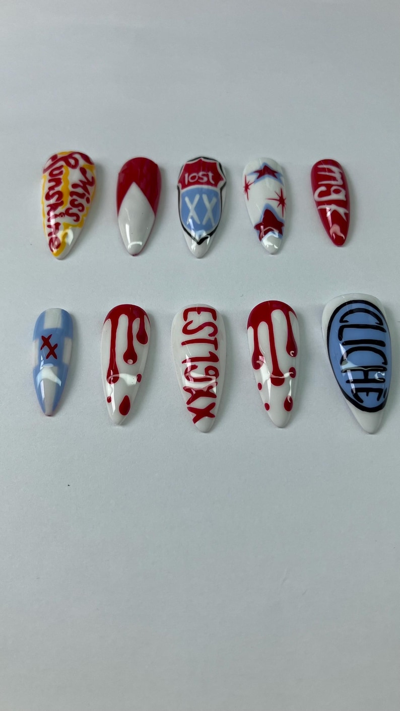 May include: A set of ten artificial nails with various designs. Some nails feature text like "Miss Sunshine" and "EST 19XX". Others have red and white patterns, including dripping blood effects and a blue and white design with the word "Lost".