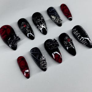 May include: A set of black and red artificial nails with various designs. Some nails feature silver embellishments, while others have a rose pattern or the name "ADELINE" in silver. The nails are oval-shaped and have a glossy finish.