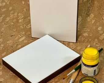 May include: Two white artist canvases with dark brown edges are displayed. A jar of yellow paint, a small dish of yellow paint, and two paintbrushes are also present. The items are on a textured, light brown surface.