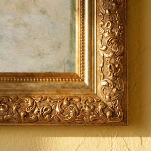 Könnte beinhalten: Nahaufnahme eines goldfarbenen, verzierten Bilderrahmens. Der Rahmen hat detaillierte Schnitzereien und eine strukturierte Oberfläche. Der Rahmen ist rechteckig mit hellem Innenraum.