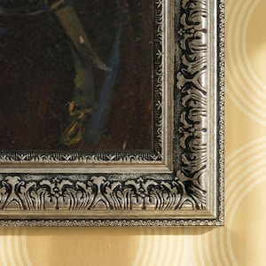 Pode incluir: Close-up de uma pintura emoldurada com uma moldura prateada ornamentada. A moldura tem entalhes detalhados e um acabamento metálico. A pintura no interior é escura com pinceladas visíveis, sugerindo uma pintura a óleo. O fundo é uma parede amarelo claro com um padrão circular.