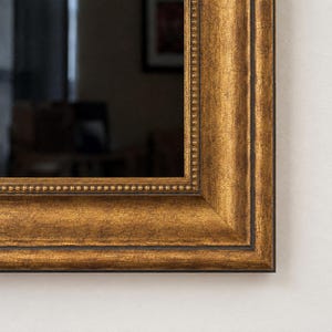 Può includere: Uno specchio da parete rettangolare con un'ampia cornice color oro anticato e una finitura scura e vissuta. Il bordo interno della cornice è decorato con una delicata fila di piccole perline in rilievo, che creano un bordo testurizzato.