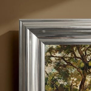 Pode incluir: Uma vista de canto de uma pintura emoldurada. A moldura é prateada com um design texturizado e canelado. A pintura retrata uma paisagem com árvores e folhagem em tons de verde e castanho.