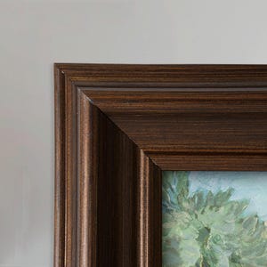 May include: A corner view of a framed painting. The frame is a dark brown with a ridged texture. The painting depicts a landscape with green trees and a light blue sky.