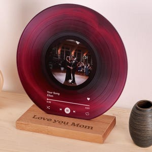 Könnte beinhalten: Eine rote Schallplatte mit einem Foto eines tanzenden Paares in der Mitte. Die Schallplatte steht auf einem Holzständer mit dem eingravierten Text "Love you Mom". Die Schallplatte hat eine digitale Musikplayer-Oberfläche mit dem Song "Your Song" von Elton John, der abgespielt wird.