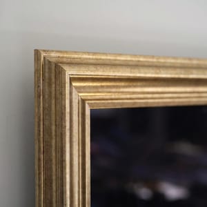 Könnte beinhalten: Nahaufnahme eines goldfarbenen rechteckigen Bilderrahmens. Der Rahmen hat ein detailliertes, geriffeltes Design und eine warme, metallische Oberfläche. Die Ecke des Rahmens ist sichtbar und zeigt die Handwerkskunst und den Stil des dekorativen Stücks.