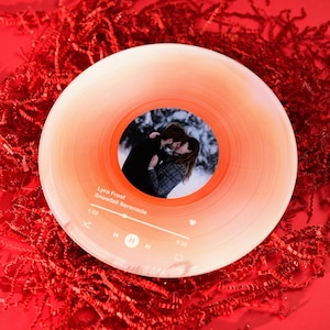 Może przedstawiać: Czerwono-biała płyta winylowa z fotografią pary całującej się w śnieżnym lesie. Płyta jest oznaczona jako "Lyra Pruitt - Snowfall Serenade".