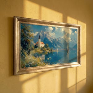 Puede incluir: Una pintura de paisaje enmarcada representa una serena escena lacustre con una iglesia, montañas y veleros. La obra de arte presenta un marco dorado y se exhibe en una pared amarilla. Los colores de la pintura incluyen azules, verdes y blancos.
