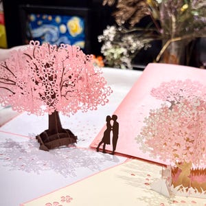 May include: A selection of pink cherry blossom pop-up cards. One card has a 3D tree, another a printed tree, and a third a 3D scene. A silhouette of a kissing couple is on one card. The cards are on a white surface.