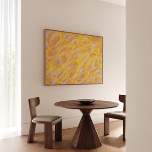 May include: A round wooden table with a conical base and two wooden chairs with beige cushions. A framed abstract painting with yellow and orange swirls hangs on the wall behind the table.