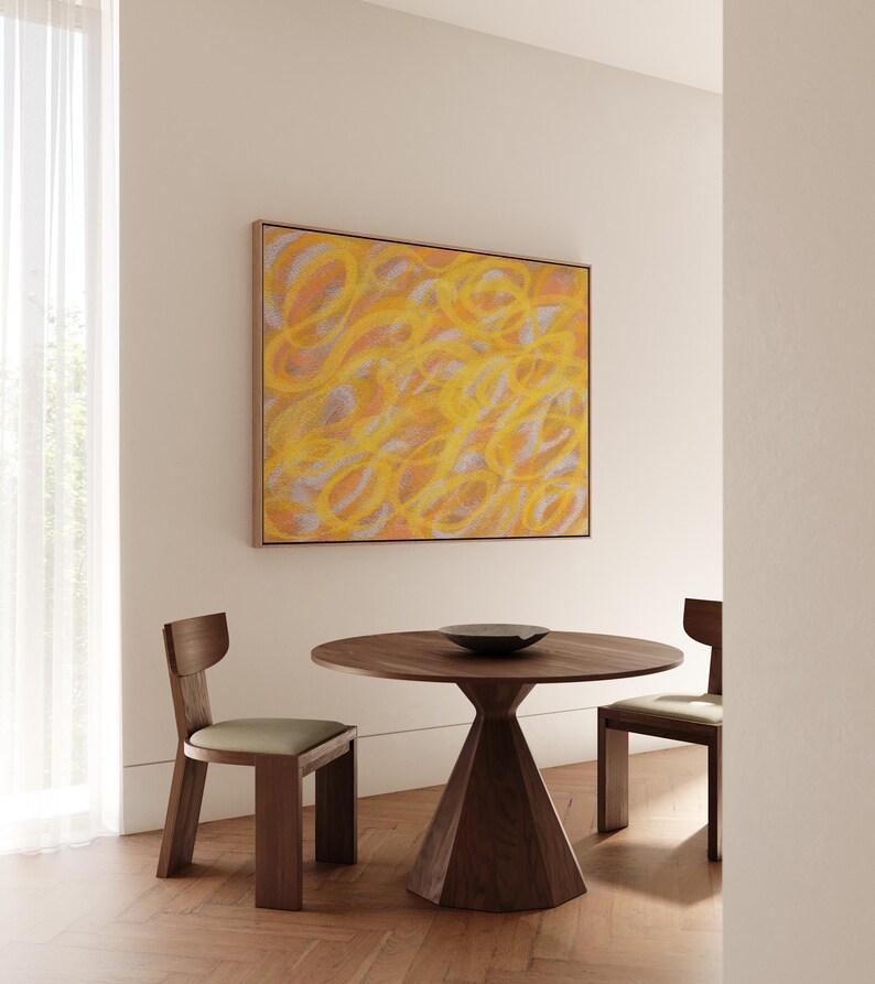 May include: A round wooden table with a conical base and two wooden chairs with beige cushions. A framed abstract painting with yellow and orange swirls hangs on the wall behind the table.