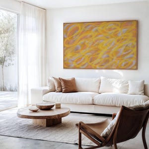 May include: A white sofa with brown throw pillows sits in a living room with a wooden coffee table and a large abstract painting with yellow and orange swirls on the wall.