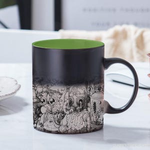 Puede incluir: Una taza de cambio de color negro y verde con un estampado gráfico blanco y negro de un diseño de criatura caprichosa. La taza tiene un interior verde y un exterior negro.