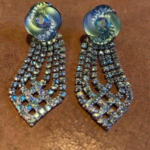 Può includere: Un paio di orecchini pendenti con un design a forma di fiore in vetro iridescente verde e blu nella parte superiore e una cascata di strass trasparenti a forma di lacrima.