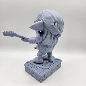 以下が含まれることがあります： ギターを弾いているゴブリンのグレーの3Dプリントフィギュア。ゴブリンはバンダナとジャケットを着ています。フィギュアはレンガの台座の上に立っています。