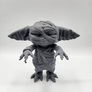 Puede incluir: Una figurita gris impresa en 3D de una criatura con apariencia de momia. La criatura tiene orejas grandes, una boca ancha y est&aacute; envuelta en vendas. Tiene tres dedos en cada pie.