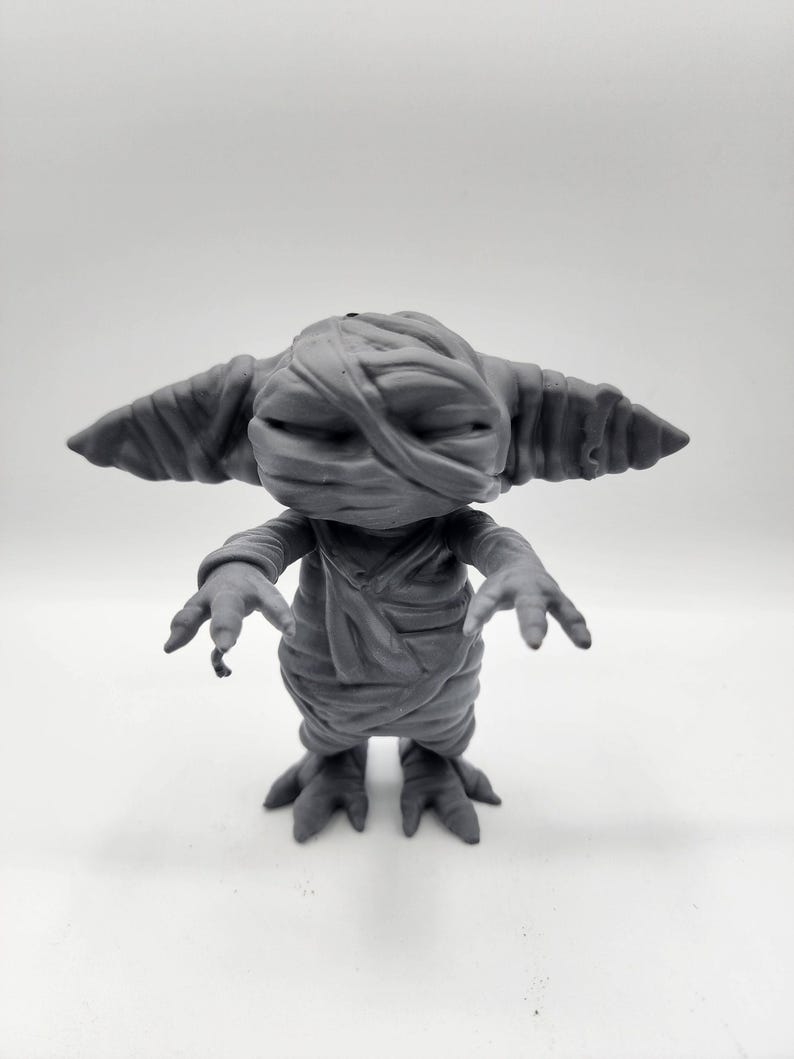 Puede incluir: Una figurita gris impresa en 3D de una criatura con apariencia de momia. La criatura tiene orejas grandes, una boca ancha y est&aacute; envuelta en vendas. Tiene tres dedos en cada pie.