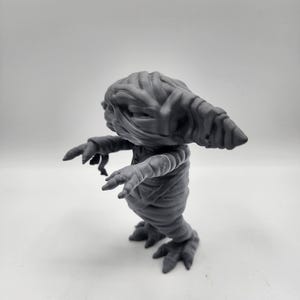 Puede incluir: Una figura gris, similar a una momia, con una cabeza larga y puntiaguda y los brazos extendidos. La figura est&aacute; hecha de un material liso, similar al pl&aacute;stico, y parece ser un juguete.