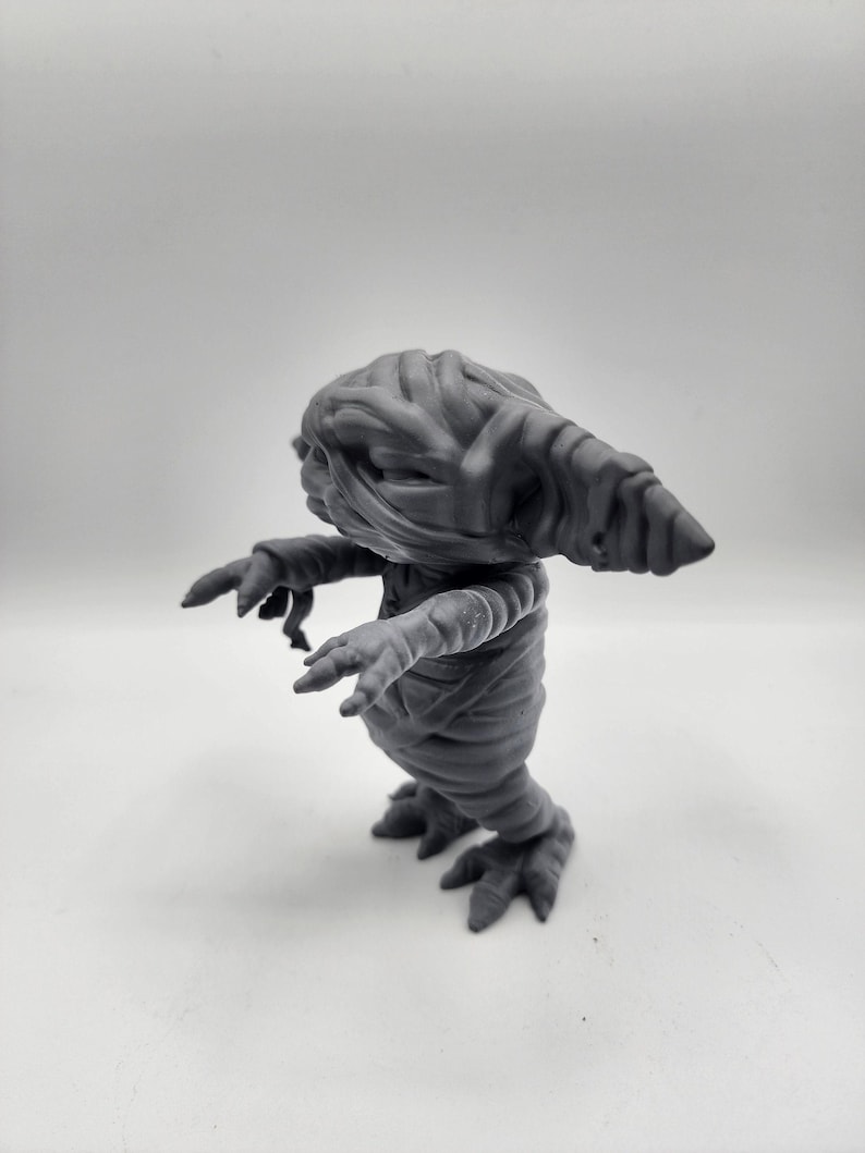 Puede incluir: Una figura gris, similar a una momia, con una cabeza larga y puntiaguda y los brazos extendidos. La figura est&aacute; hecha de un material liso, similar al pl&aacute;stico, y parece ser un juguete.